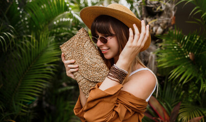 Close up summer fashionable  portrait of brunette woman in straw hat posing on tropical palm leaves background in Bali. Wearing stylish bohemian accessories.