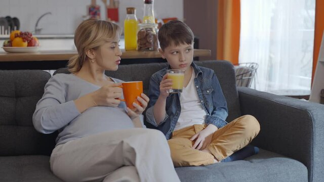Pregnant woman is chatting while drinking coffee or tea, her little son is drinking juice.Pregnancy, motherhood concept. Slow motion video.