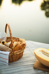 Fototapeta premium A beautiful picnic for two by the lake. A magical sunset over the lake. A basket of bread, glasses and a melon on a blanket. A romantic date for two.