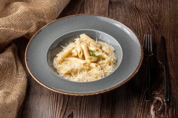 Pasta Penne with parmesan cheese and creamy sauce. Isolated dish in a grey round plate on wooden background. Sacking and silver cutlery by side. Mediterranean gourmet food.
