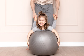 Father and daughter are training and playing at home. Smiling, cute, funny, happy, amotional, adorible child in casual clothes doing exercises with grey fit gymnastic ball. healthy concept