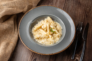 Pasta Penne with parmesan cheese and creamy sauce. Isolated dish in a grey round plate on wooden background. Sacking and silver cutlery by side. Mediterranean gourmet food. Top view.