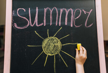 A little girl beautifully, diligently writes on a wooden board, easel, holding yellow chalk in her hand, the word summer and draws the sun. Child's dream of vacation.