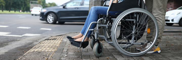 Female legs in wheelchair close up. Help for people with physical disabilities. Man drive wheelchair and stand at pedestrian crossing. Social services for population.