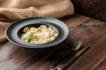 Pasta Farfalle with parmesan cheese and creamy sauce. Isolated dish in a black round plate on wooden background. Sacking and silver cutlery by side. Mediterranean gourmet food.
