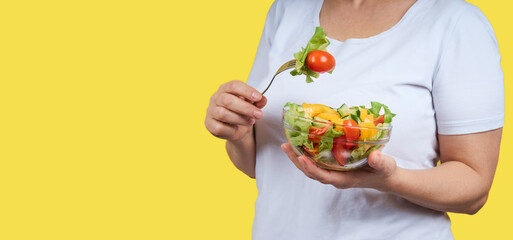 Cropped image of woman in white shirt holding a plate of vegetable salad. Studio shot on yellow background with copy space.