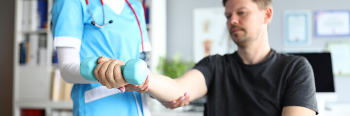 Doctor feel hand in hospital. Man sit on chair with his arm extended forward with dumbbell at doctor appointment. Woman in doctor suit and red stethoscope examine visitor hand for fracture