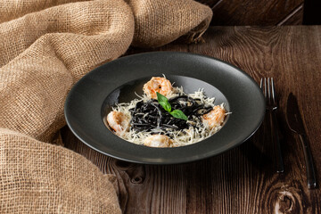 Black spaghetti. Black pasta with shrimps and parmesan in a black round plate isolated on wooden background. Silver cutlery and sacking by side. Mediterranean gourmet food.