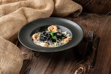 Black spaghetti. Black pasta with shrimps and parmesan in a black round plate isolated on wooden background. Silver cutlery and sacking by side. Mediterranean gourmet food.