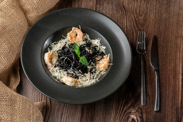 Black spaghetti. Black pasta with shrimps and parmesan in a black round plate isolated on wooden background. Silver cutlery and sacking by side. Mediterranean gourmet food. Top view.