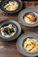 Four kinds of different cooked pastas: Penne, Farfalle, Carbonara, Black Spaghetti. Close-up isolated on wooden background in black and grey plates. Mediterranean gourmet food. Top view.