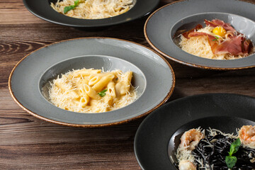 Four kinds of different cooked pastas: Penne, Farfalle, Carbonara, Black Spaghetti. Close-up isolated on wooden background in black and grey plates. Mediterranean gourmet food.