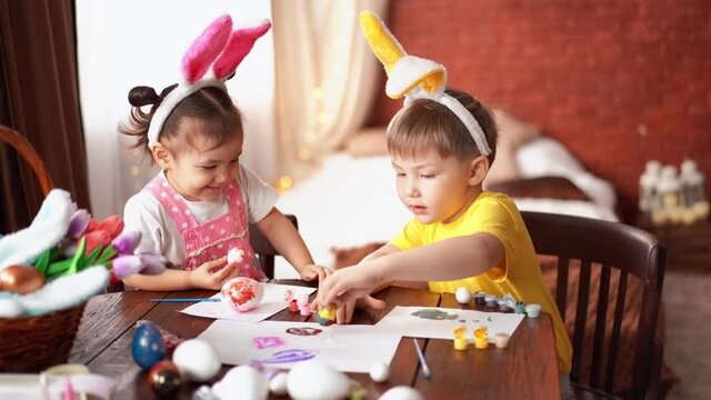 Brother and sister are painting Easter eggs for Easter. Happy children paint eggs in a playful way. Children have rabbit ears on their heads.