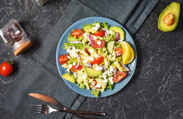 Vegetarian healthy lunch - pasta salad with fresh vegetables, avocado and feta on a black stone background