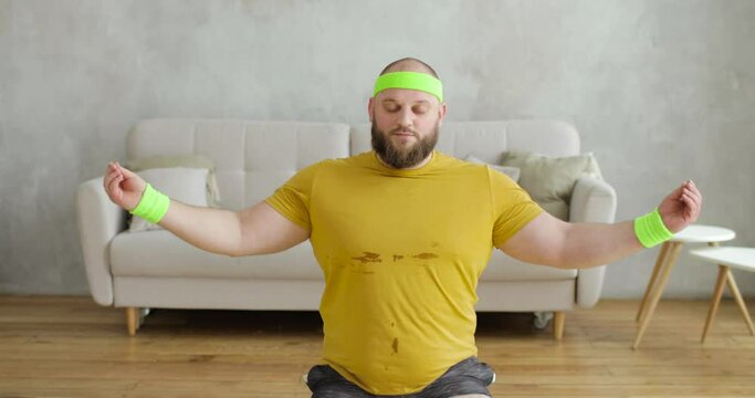 Funny fat man newcomer in sport in yellow sportswear is doing meditation sitting on yoga mat in living room. Fitness exercises at home. Joke, mem, humor, parody. Workout, training concept.
