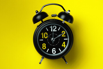 A black alarm clock with large numbers lies on a yellow background. The concept of the time,...