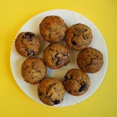 eight muffins with cherries, small pieces of dark chocolate, rice flour, corn flour, eggs and yogurt lie on a white plate on a yellow background top view . homemade gluten-free cupcakes