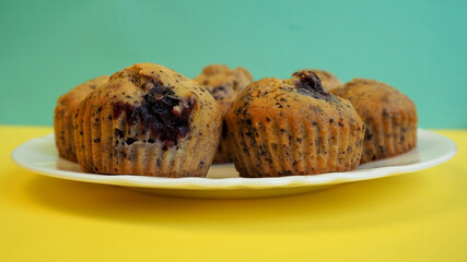 six muffins with cherries, small pieces of dark chocolate, rice flour, corn flour, eggs and yogurt lie on a white plate on a yellow with a green background side view.  gluten free food