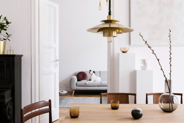 Stylish scandinavian interior of living room with design wooden table, chairs, grey sofa, decoration, personal accessories and beautiful dog in elegant home decor.