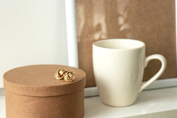 Still life. Cup of coffee, blank beige poster frame, craft round box and beautiful gold earrings on the white wall. Side view