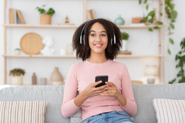 Mixed race woman in headphones holds smartphone, resting on couch, spend lazy weekend