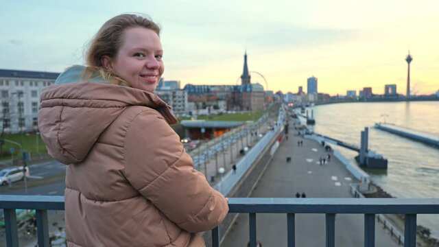 [4k] young blonde woman in winter coat turning to camera in front of european city at river rhein