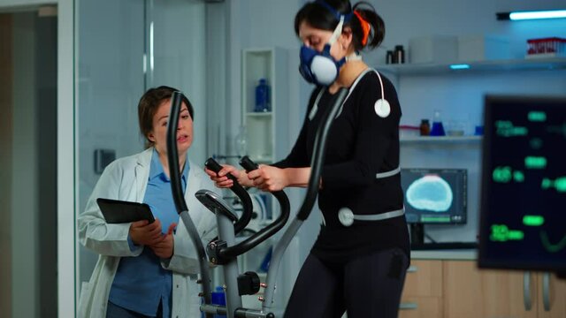Woman athlete with mask running on cross trainer in sports science lab measuring performance and oxygen consumption during Vo2max test. Medical researcher looking at EKG scan monitoring heart rate