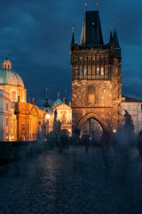 Fototapeta premium Blurred ghost like people silhouettes walking on illuminated Charles Bridge in Prague in night on Gothic towers background
