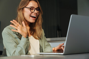 Happy nice woman waving hand and winking while working with laptop