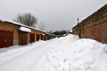 Obraz na płótnie Canvas A cloudy winter day. Snowfall. Rows of one-story brick garages with closed metal painted gates. Drifts of snow near the walls along the road. Snow on the roofs.