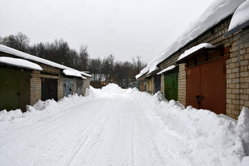A cloudy winter day. Snowfall. Rows of one-story brick garages with closed metal painted gates. Drifts of snow near the walls along the road. Snow on the roofs.