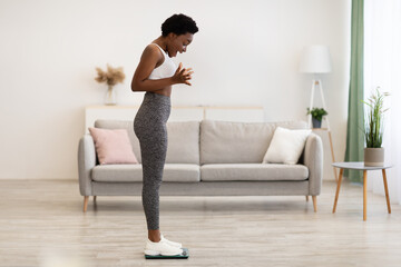 Fototapeta na wymiar Joyful Black Lady Weighing Standing On Weight-Scales At Home
