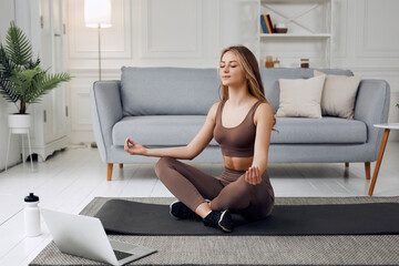 Young healthy beautiful woman in sportive top and leggings practicing yoga at home sitting in lotus...