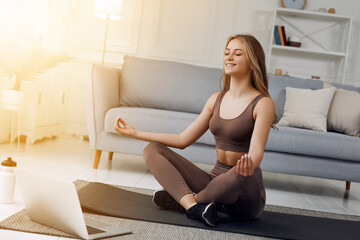 Young healthy beautiful woman in sportive top and leggings practicing yoga at home sitting in lotus pose on yoga mat meditating smiling relaxed with closed eyes, Mindfulness meditation concept