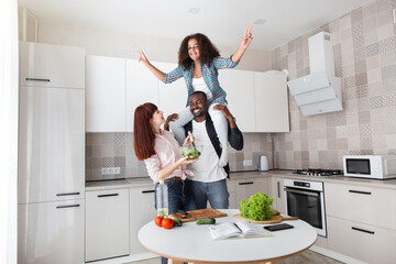 Enjoy communication and cookery hobby concept. Happy black man dad holding his curly daughter at the shoulders and lookiing at his pretty caucasian wife with bowl with salad