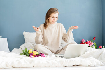 Obraz na płótnie Canvas Happy woman sitting on the bed wearing pajamas holding tulip flowers bouquet chatting on laptop