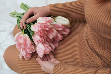 Pink tulips in the hands of a woman. A woman sits on the bed and holds a bouquet on her lap.