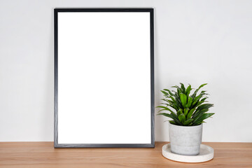 Black wooden vertical frame with white blank card and green plant in concrete pot on wooden table on light gray wall background. Mockup, template for your design, free copy space for text.