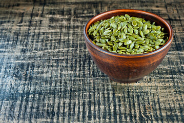 Peeled pumpkin seeds in a clay bowl on a wooden table. Seeds on a black shabby board. Copy space and free space for text near the seeds.