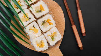 Dessert sushi. Sweet kiwi, pineapple sushi rolls. Sushi on a wooden tray on a black background with a tropical leaf