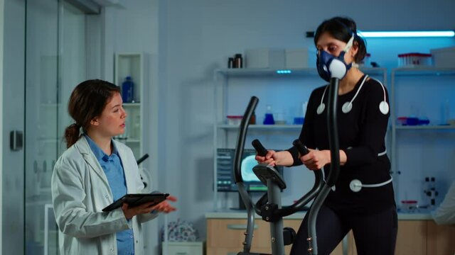 Woman with mask running on cross trainer testing heart rate using electrodes, medical researcher writing EKG results on tablet. Performance athlete monitoring physical endurance in science sport lab