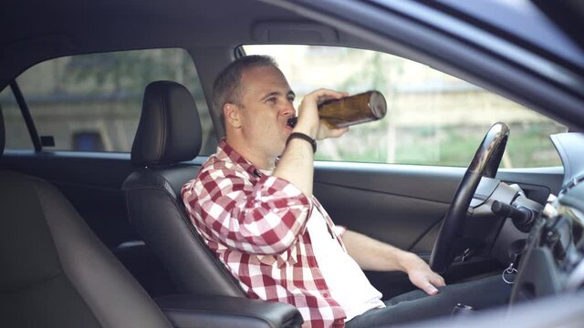 Careless Caucasian adult man drinking beer sitting on driver's seat in car. Side view portrait of frivolous grey-haired middle aged guy abusing alcohol driving vehicle. Alcoholic lifestyle.