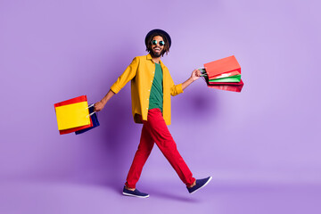 Full length photo of sweet cute dark skin guy dressed yellow shirt walking holding shopping bags isolated purple color background