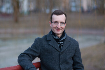 Portrait of a man with glasses and a coat. He sits on a bench in an autumn city park.