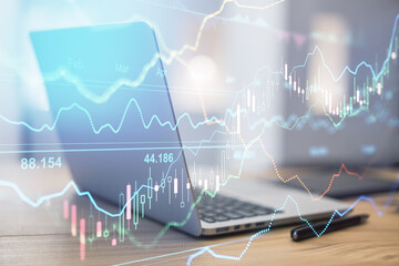 Online stock market concept with transparent digital screen with financial graphs and opened laptop on wooden table at background