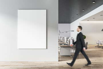 Businessman goes to blank white poster on light grey wall in modern open space office with light furniture and wooden floor