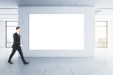 Businessman in black suit goes by blank white poster on the wall in the center of empty hall with city view
