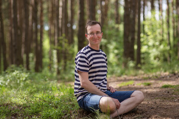 Portrait of a smiling man with glasses. He sits on the ground in a bright sunny spring forest.