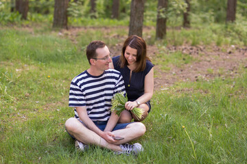 Naklejka na ściany i meble Man and woman sit on the grass in a bright spring forest. The man is holding a smartphone, the woman is holding a fresh bouquet of lilies of the valley.