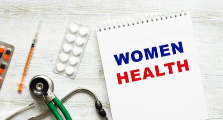 WOMEN HEALTH is written in a notebook on a white table next to pills and a stethoscope.